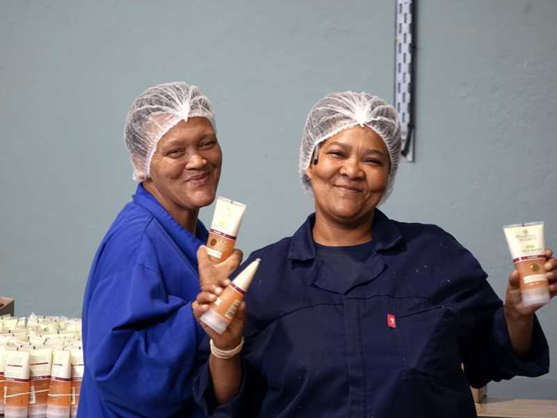 Zwei lächelnde Mitarbeiterinnen von African Extracts halten fertige Hautpflegeprodukte in den Händen.