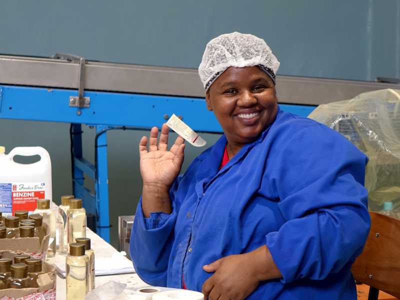Smiling employee at African Extracts holding a label on her finger in the packaging area.